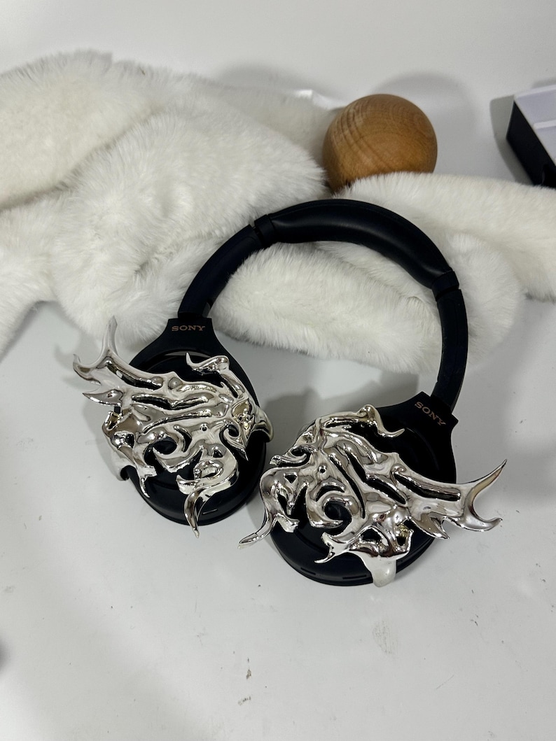 May include: Black Sony headphones with silver decorative accents. The headphones have a black headband and earcups. The silver accents are an ornate, flame-like design. The headphones are on a white surface.