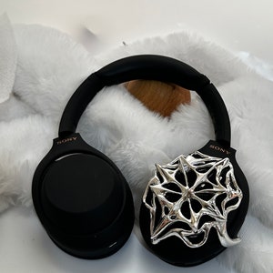 May include: Black Sony headphones with a silver, star-shaped decorative accent on one ear cup. The headphones have a padded headband and earcups. The background is a white, fluffy surface.