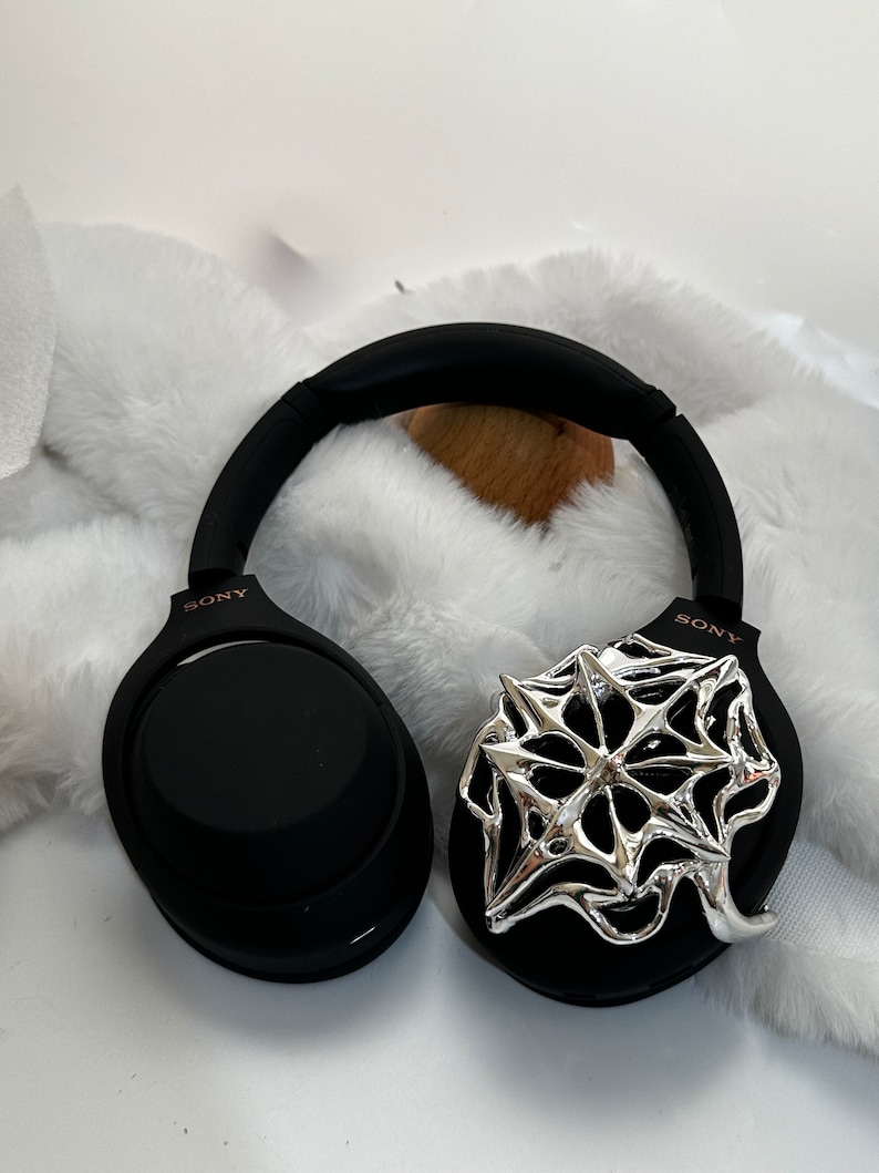 May include: Black Sony headphones with a silver, star-shaped decorative accent on one ear cup. The headphones have a padded headband and earcups. The background is a white, fluffy surface.
