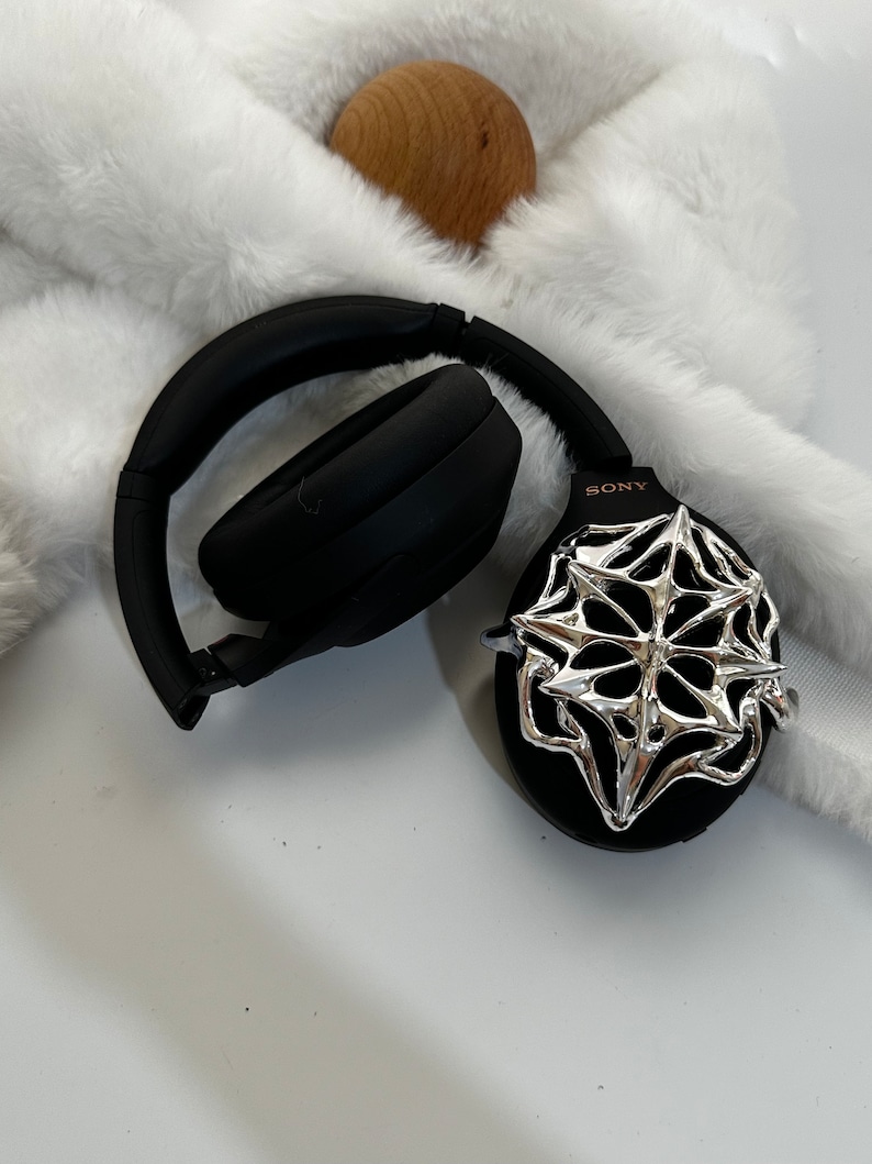 May include: Black Sony headphones with a silver decorative accent. The headphones are resting on a white, fluffy surface. A small wooden sphere is also visible in the image.