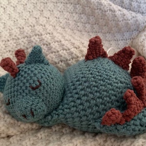 Könnte beinhalten: Ein gehäkeltes Plüschtier in Form eines schlafenden Drachen. Der Drache ist blaugrün mit braunen Hörnern und Stacheln. Der Drache liegt auf der Seite mit dem Kopf auf seinen Vorderpfoten.