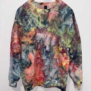 May include: A tie-dye sweatshirt in a variety of colors, including pink, yellow, green, and blue. The sweatshirt is hanging on a wooden hanger against a white background. The garment has a crew neck and long sleeves.