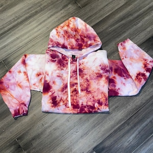 May include: A tie-dye hoodie in shades of burgundy, white, and peach. The hooded sweatshirt has long sleeves and a drawstring at the neckline. The fabric has a marbled effect, with the colors blending together. The hoodie is laying flat.