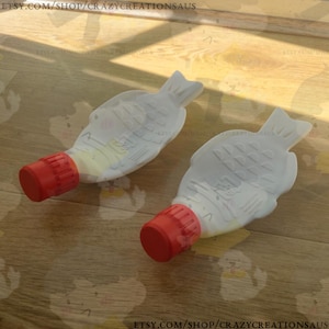 May include: Two white fish-shaped condiment bottles with red caps. The bottles are made of plastic and have a textured surface. The bottles are on a wooden surface.