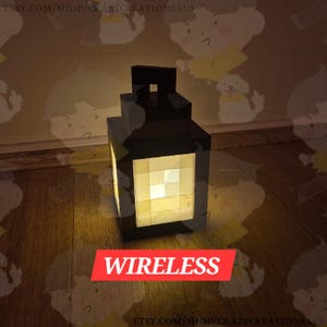 May include: A black, cube-shaped lantern with a bright, yellow-toned light emanating from its center. The word "WIRELESS" is displayed in a red banner at the bottom. The lantern is sitting on a wooden surface.