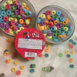 May include: Two clear bowls filled with colorful, ring-shaped cereal, one with milk and a spoon. A red container of Kellogg's Froot Loops cereal is in the foreground. The cereal is scattered on a white surface.