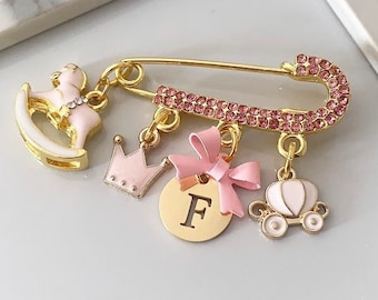 Può includere: Spilla da balia dorata con strass rosa. Presenta ciondoli pendenti: un cavallo a dondolo, una corona, un disco con la lettera "F", un fiocco e una carrozza. I ciondoli sono rosa e bianchi.