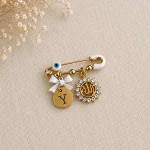 May include: A gold-tone safety pin brooch featuring a white enamel evil eye bead, a small white bow, a circular gold charm engraved with the letter Y, and a circular gold charm with Arabic calligraphy surrounded by clear rhinestones.
