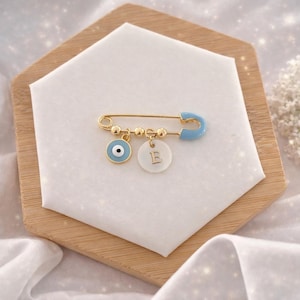 May include: Gold-coloured safety pin with a light blue enamel clasp. The pin features a small blue and white evil eye charm and a white disc charm with the letter "E". The pin is displayed on a white surface.