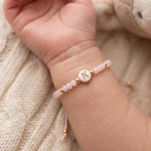 Peut inclure: Un bracelet de perles roses avec un charm circulaire blanc portant la lettre "K" en or. Le bracelet comporte de petites perles dorées et est porté au poignet d'un bébé.