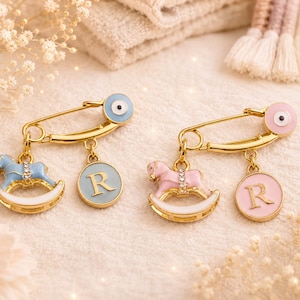 May include: Two gold-tone safety pins featuring an evil eye charm, a rocking horse charm with clear crystals, and a circular charm with the letter R. One pin has light blue enamel accents, and the other has light pink enamel accents.