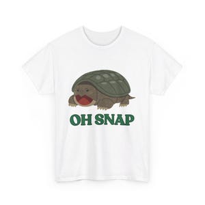 以下が含まれることがあります： 緑色の「OH SNAP」の文字が書かれた、白いTシャツに、漫画のミシシッピアカミミガメのイラストが描かれています。