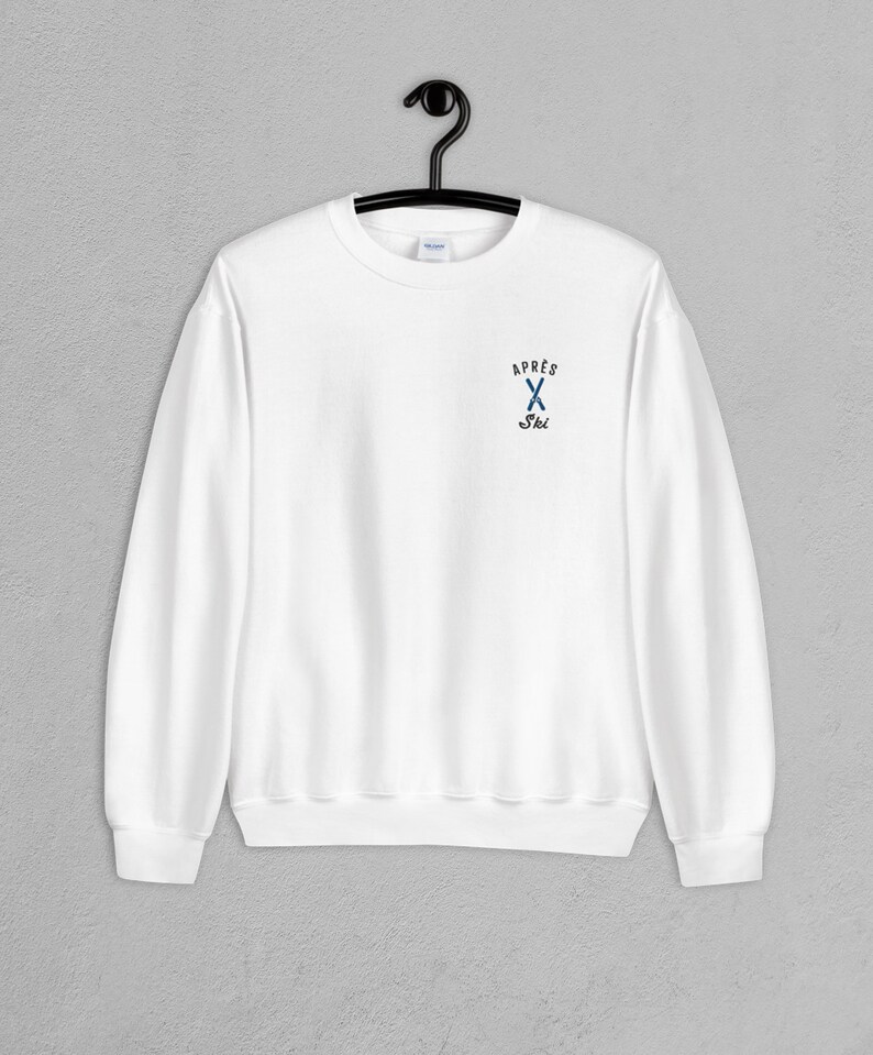 Puede incluir: Sudadera blanca de cuello redondo con un peque&ntilde;o logotipo bordado en el pecho izquierdo. El logotipo es azul y negro y dice "Apres Ski" con un par de esqu&iacute;s cruzados.