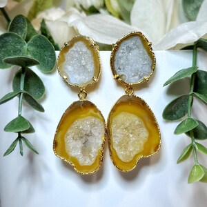 Puede incluir: Pendientes dorados con rodajas de geoda natural. Los pendientes tienen una combinación de colores amarillo y blanco con un borde dorado. Los pendientes cuelgan frente a un jarrón blanco con vegetación.