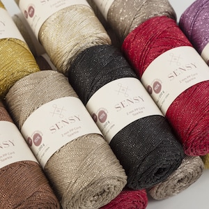 Peut inclure: Gros plan sur six bobines de fil brillant en cordon PP de 2 mm dans diff&eacute;rentes couleurs, dont l'or, le beige, le noir, le rouge et le violet. Chaque bobine porte une &eacute;tiquette avec le nom de la marque "SENSY" et le texte "90% / 200 g / 251/230m".