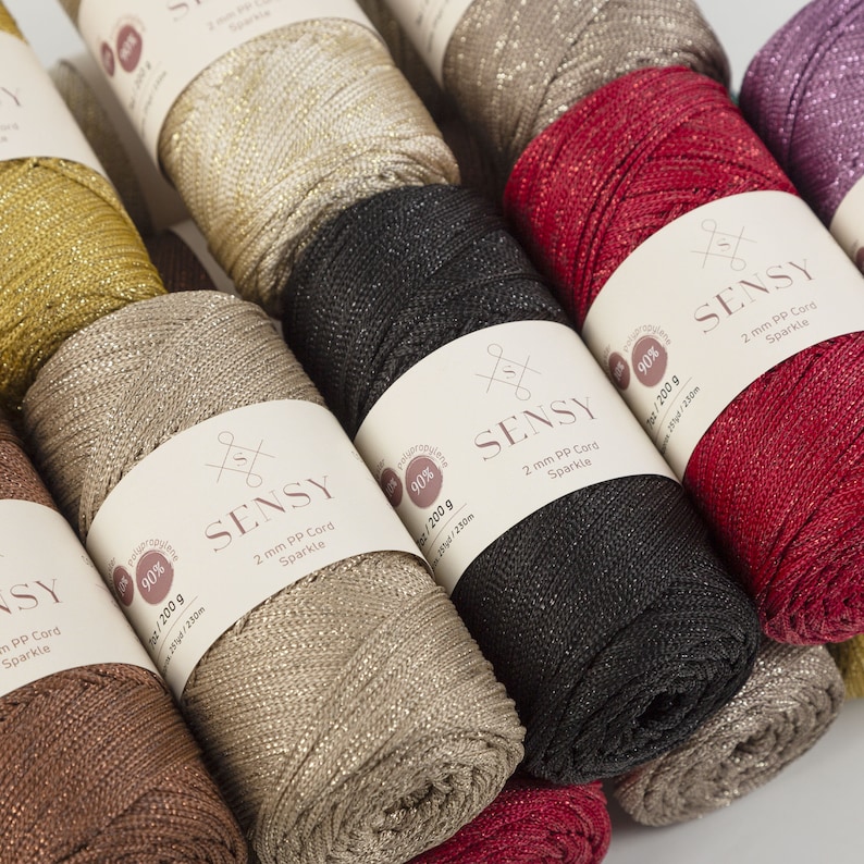 Peut inclure: Gros plan sur six bobines de fil brillant en cordon PP de 2 mm dans diff&eacute;rentes couleurs, dont l'or, le beige, le noir, le rouge et le violet. Chaque bobine porte une &eacute;tiquette avec le nom de la marque "SENSY" et le texte "90% / 200 g / 251/230m".