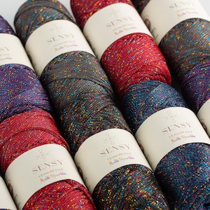 May include: Close-up of several spools of Sensy Multi Sparkle yarn in various colors, including red, purple, and teal. Each spool has a white label with the Sensy logo and product details. The yarn is tightly wound and has a sparkly texture.