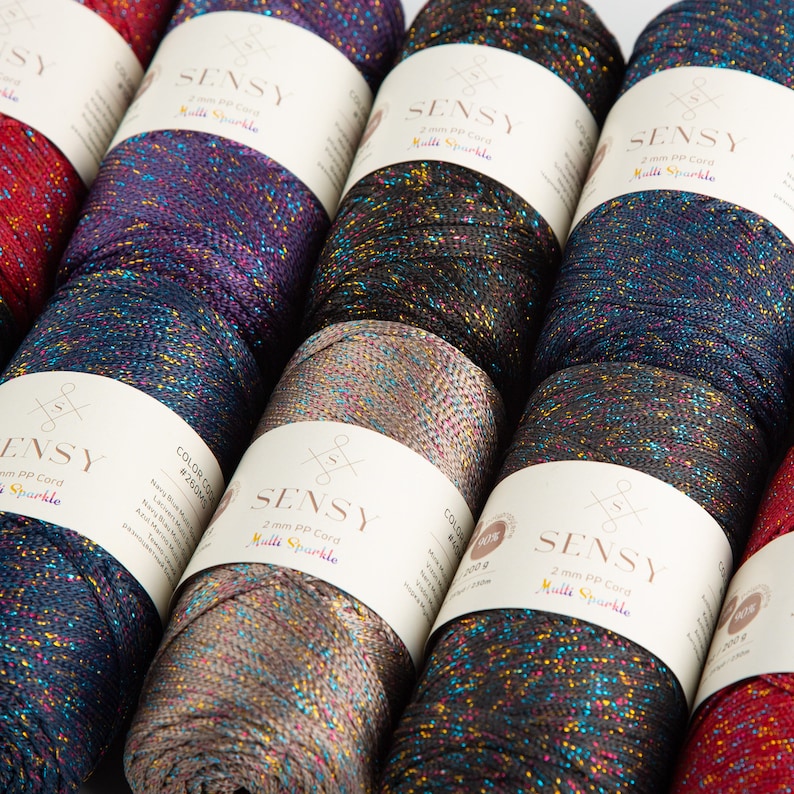 Puede incluir: Primer plano de diez carretes de hilo de colores brillantes. El hilo es un cord&oacute;n PP de 2 mm y est&aacute; etiquetado como "SENSY Multi Sparkle".