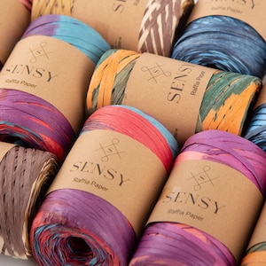 Può includere: Assortimento di rotoli di rafia di carta colorati con etichette marroni che recitano "Sensy Raffia Paper".