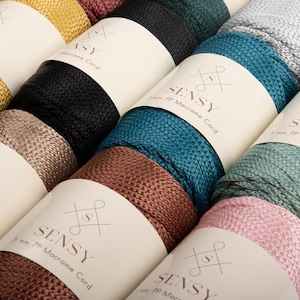 May include: Close-up of several spools of Sensy 5mm PP Macrame Cord in various colors, including black, teal, brown, and pink. Each spool is labeled with the Sensy logo and product information. The macrame cord is tightly wound, showcasing its texture and color variations.