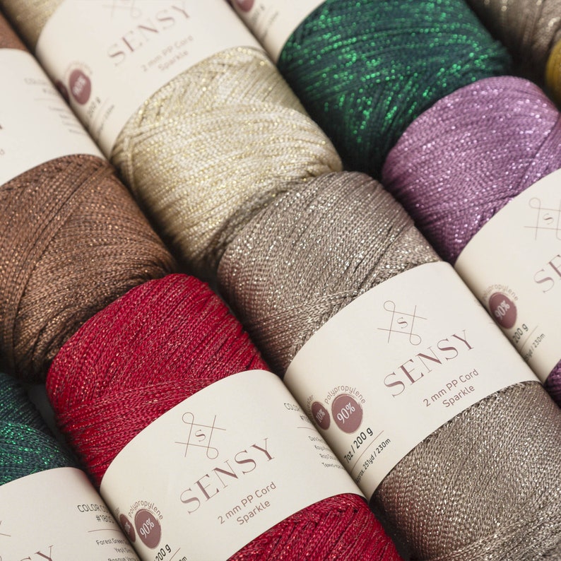 Peut inclure: Gros plan sur des bobines de cordon paillet&eacute; de diff&eacute;rentes couleurs, dont l'or, le vert, le violet, le marron et le rouge. Les bobines sont &eacute;tiquet&eacute;es "SENSY 2 mm PP Cord Sparkle 90% Polypropyl&egrave;ne 200 g 25/230m".