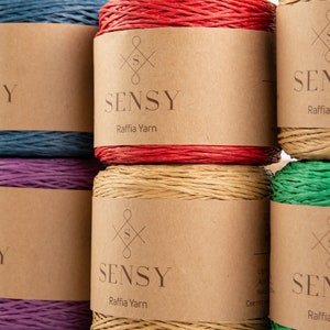 May include: Close-up of a stack of colorful raffia yarn spools. Each spool has a brown paper label with the text "SENSY Raffia Yarn".