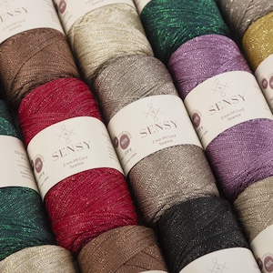 May include: Close-up of several spools of Sensy 2 mm PP Cord Sparkle yarn in various colors, including red, gold, green, and purple. Each spool has a white label with the Sensy logo and product information. The yarn appears to have a metallic sheen.