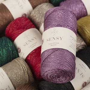 Peut inclure: Gros plan d'une bobine de cordon paillet&eacute; violet avec une &eacute;tiquette blanche indiquant "SENSY 2 mm PP Cord Sparkle". L'&eacute;tiquette indique &eacute;galement le code couleur "R7715".
