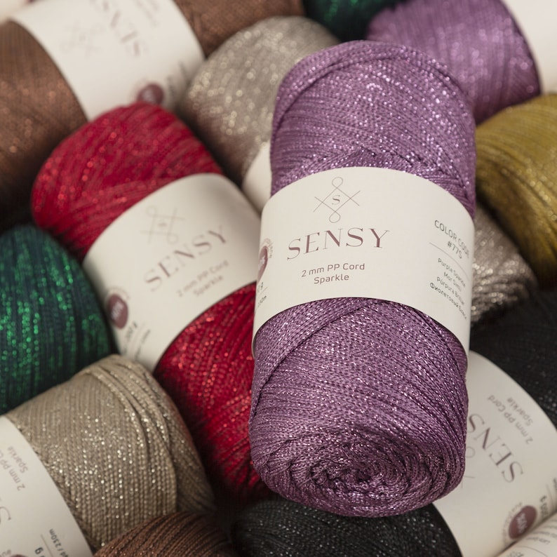 Peut inclure: Gros plan d'une bobine de cordon paillet&eacute; violet avec une &eacute;tiquette blanche indiquant "SENSY 2 mm PP Cord Sparkle". L'&eacute;tiquette indique &eacute;galement le code couleur "R7715".