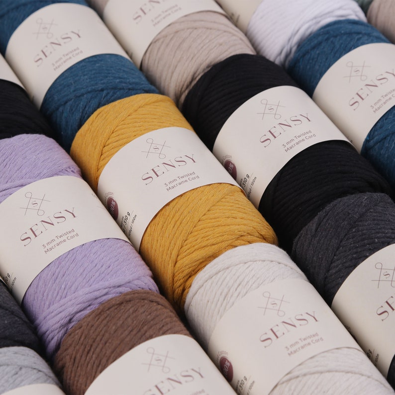 May include: A close-up of various spools of 3 mm twisted macrame cord in different colors, including mustard yellow, lavender, white, black, gray, and brown. The spools are labeled "SENSY 3 mm Twisted Macrame Cord".