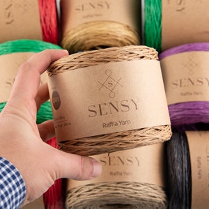 May include: A close-up of a brown raffia yarn ball with a label that reads "SENSY Raffia Yarn". The yarn is 100% natural and comes in a 250 gram ball.