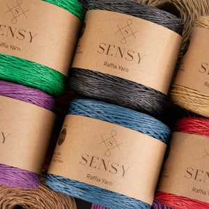 May include: Close-up of six spools of raffia yarn in various colors, including green, purple, blue, red, gray, and natural. Each spool has a brown paper label with the brand name "SENSY Raffia Yarn" printed on it.