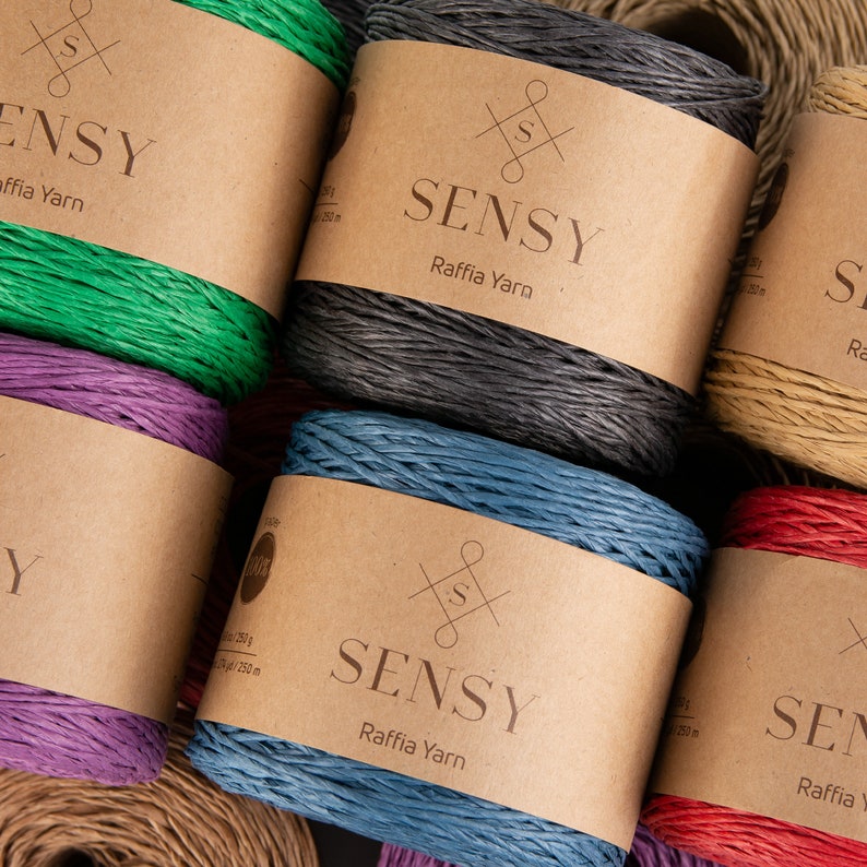 May include: Close-up of six spools of raffia yarn in various colors, including green, purple, blue, red, gray, and natural. Each spool has a brown paper label with the brand name "SENSY Raffia Yarn" printed on it.