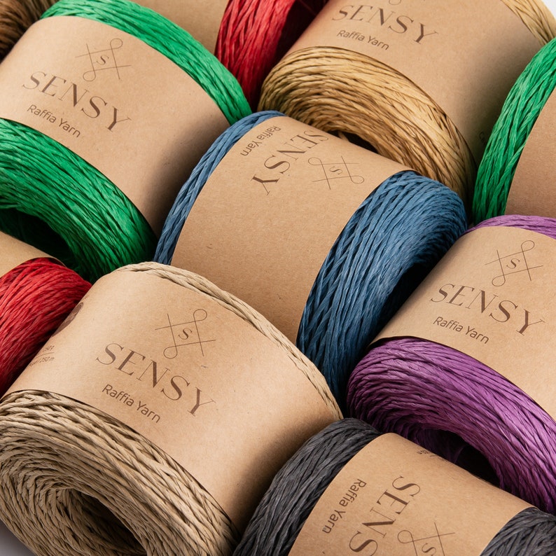 May include: Assortment of colorful raffia yarn spools with the brand name "SENSY" printed on each label. The yarn is available in green, red, beige, blue, purple, and black.