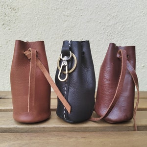Könnte beinhalten: Drei kleine Ledertaschen mit Kordelzug und einem Schlüsselring aus Metall. Die Taschen sind braun, schwarz und dunkelbraun.