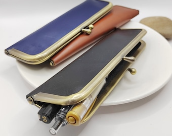 May include: Two leather pencil cases with a vintage brass clasp. One case is navy blue and the other is black. Both cases are open and have pens inside.