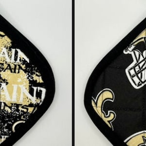 May include: Two diamond-shaped pot holders. One features a gold, black, and white Saints logo pattern. The other has a black background with a gold Saints helmet and fleur-de-lis design. Both have a black border.