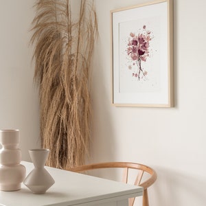 May include: A framed watercolor painting of a bouquet of pink flowers with white splatters on a white background. The painting is hanging on a white wall next to a dried pampas grass plant. A white table with a wooden chair is in the foreground.