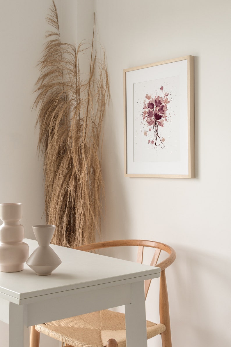 May include: A framed watercolor painting of a bouquet of pink flowers with white splatters on a white background. The painting is hanging on a white wall next to a dried pampas grass plant. A white table with a wooden chair is in the foreground.