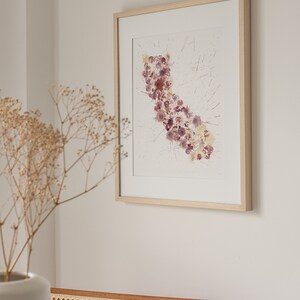 May include: A framed watercolor painting of the state of California, depicted with a cluster of pink and purple watercolor dots. The painting is framed in a light wood frame and hangs on a white wall.