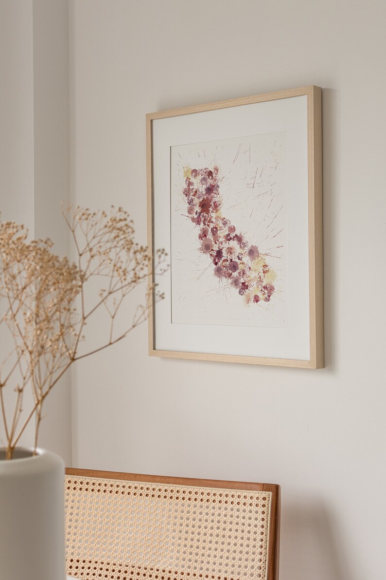 May include: A framed watercolor painting of the state of California, depicted with a cluster of pink and purple watercolor dots. The painting is framed in a light wood frame and hangs on a white wall.
