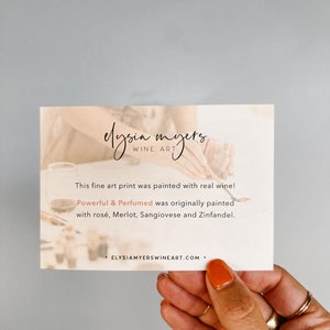 May include: A white card with the text "elysia myers, WINE ART" and the text "This fine art print was painted with real wine! Powerful & Perfumed was originally painted with ros&eacute;, Merlot, Sangiovese and Zinfandel. ELYSIAMYERSWINEART.COM."  The card is being held by a hand with orange nail polish.