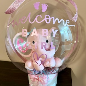 Puede incluir: Un globo de burbujas transparente con un elefante de peluche rosa en el interior, con las palabras "welcome BABY GIRL" en escritura rosa. El globo está decorado con un lazo y una cinta rosas, y una pequeña caja blanca en la parte inferior.