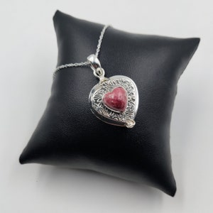 Puede incluir: Un collar con un colgante en forma de corazón de plata con una piedra preciosa rosa en el centro. El colgante tiene intrincados diseños florales alrededor de la piedra preciosa.