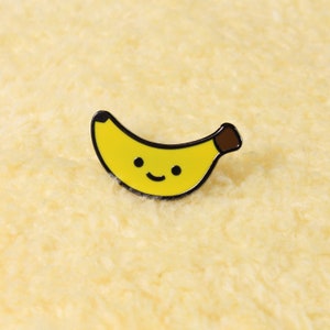 Könnte beinhalten: Gelber Emaille-Anstecker in Form einer lächelnden Banane. Der Anstecker hat eine schwarze Umrandung und einen braunen Stiel. Das Gesicht der Banane hat zwei schwarze Punkte als Augen und einen gebogenen Mund.