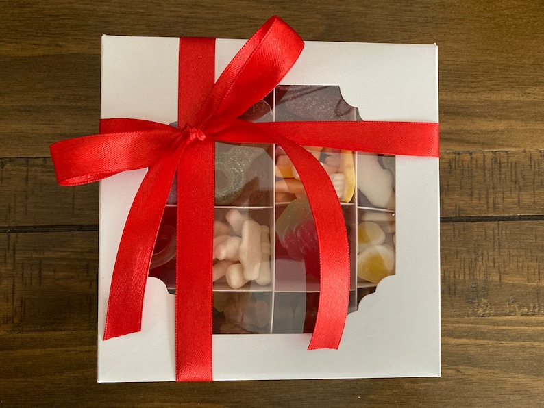 May include: A white gift box with a red ribbon tied around it. The box has a clear window on the front, revealing a variety of colorful gummy candies inside.