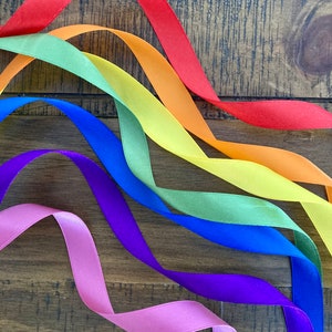 May include: A rainbow of satin ribbon in red, orange, yellow, green, blue, purple, and pink, arranged in a wave pattern on a wooden surface.