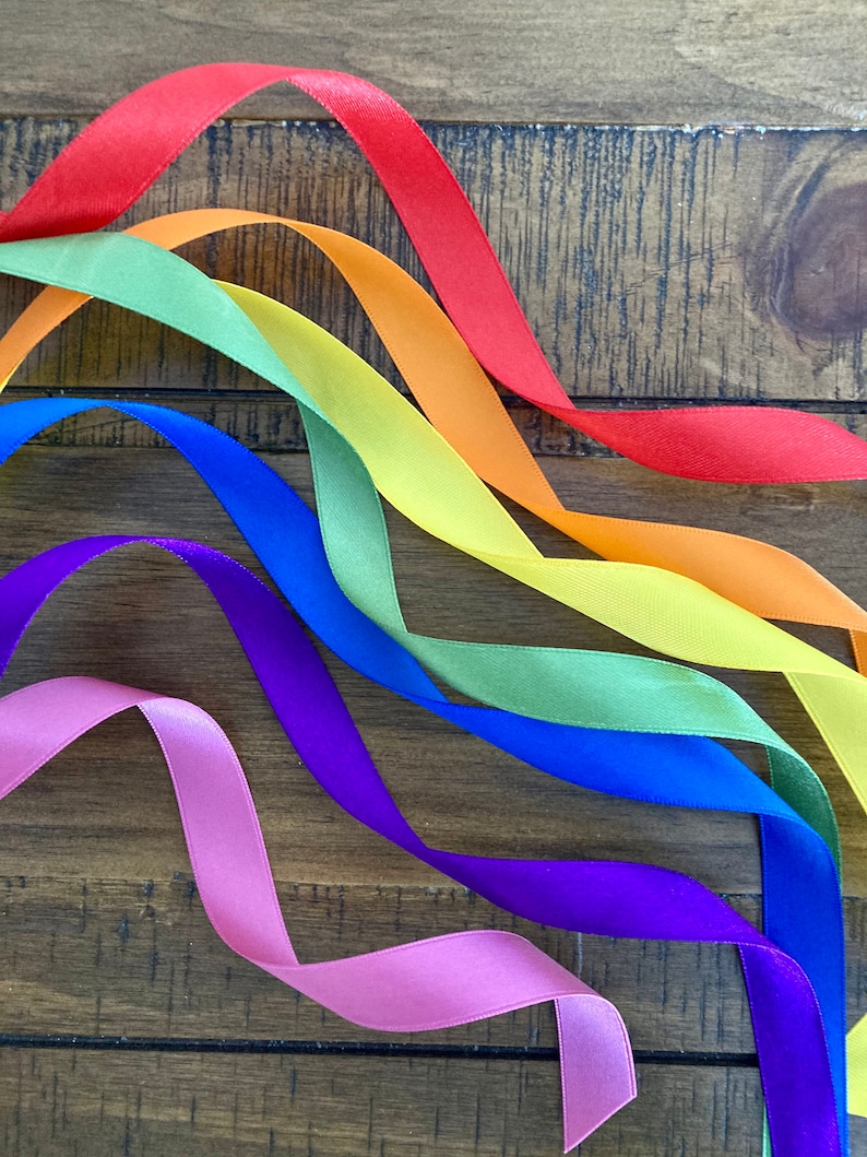 May include: A rainbow of satin ribbon in red, orange, yellow, green, blue, purple, and pink, arranged in a wave pattern on a wooden surface.