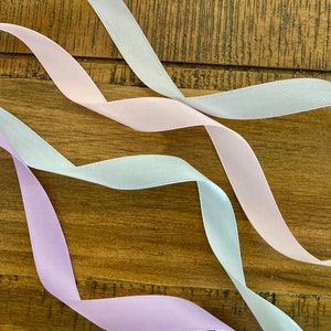 May include: Three satin ribbons in different shades of pink, light blue, and lavender, arranged in a wavy pattern on a wooden surface.