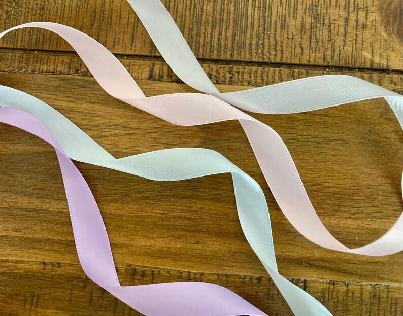 May include: Three satin ribbons in different shades of pink, light blue, and lavender, arranged in a wavy pattern on a wooden surface.